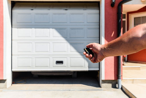 A hand holding a garage door opener remote while a garage door opens in the background.